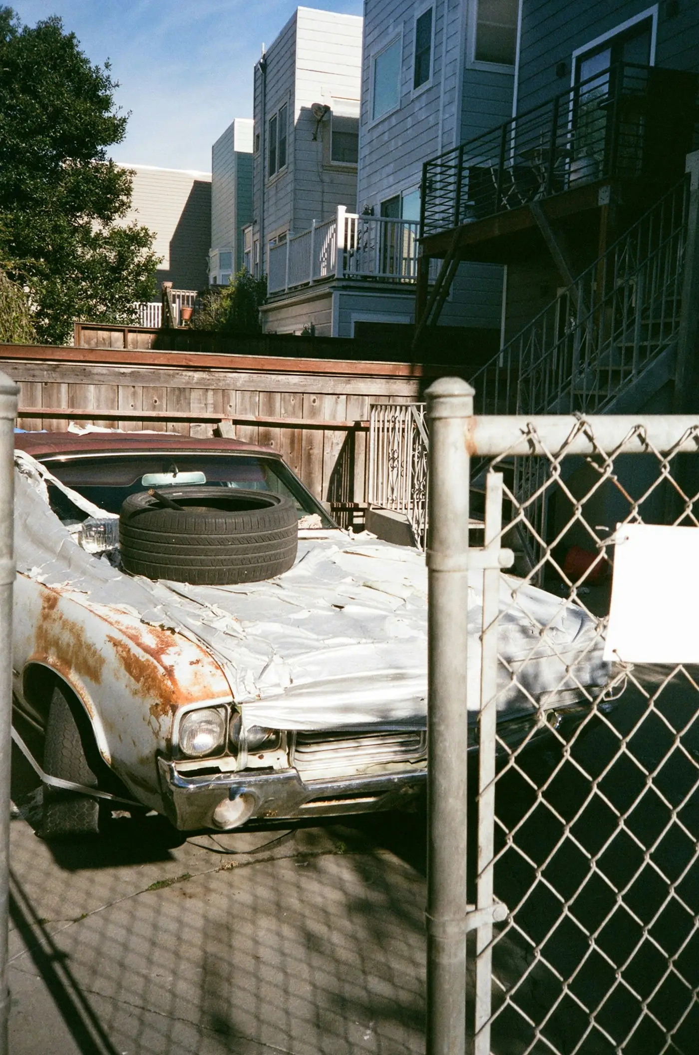 An old rusted sedan with a spare tire on its hood, parked behind a chain-link gate